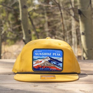 Puede incluir: Gorra de color amarillo mostaza con visera plana y detalle de cuerda negra. La gorra presenta un parche rectangular con una escena de montaña y el texto "SUNSHINE PEAK" y "SAN JUAN RANGE". La gorra está sobre una superficie de madera.