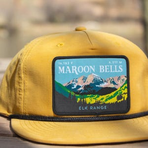 Könnte beinhalten: Senfgelbe Baseballkappe mit schwarzem Seildetail und einem rechteckigen Aufnäher. Der Aufnäher zeigt eine Berglandschaft mit dem Text "MAROON BELLS" und "ELK RANGE".
