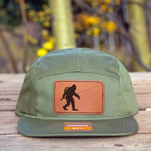 Könnte beinhalten: Olivgrüne Fünf-Panel-Kappe mit einem braunen Lederaufnäher mit einer schwarzen Silhouette von Bigfoot mit einer Angelrute. Die Kappe hat einen flachen Schirm und ein kleines Etikett unten.