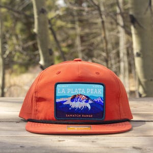 May include: An orange baseball cap with a flat brim and a black rope detail across the base of the crown. A rectangular patch on the front features a graphic of La Plata Peak in the Sawatch Range with blue, white, and dark purple colors.