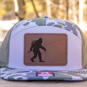 Puede incluir: Una gorra de camionero de camuflaje blanco y verde con un parche de cuero marrón que presenta una silueta de Bigfoot sosteniendo una caña de pescar. La gorra tiene una parte trasera de malla y un panel frontal blanco.