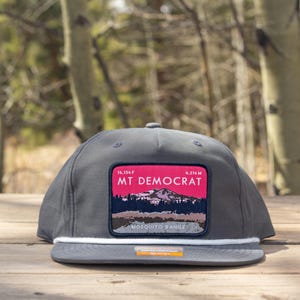 May include: A grey baseball cap with a flat peak and a white rope detail. The cap has a rectangular patch with the text "MT DEMOCRAT" and a mountain range graphic. The patch is outlined in white with a pink background.