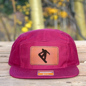 Könnte beinhalten: Magentafarbene Fünf-Panel-Kappe mit einem braunen Lederpatch, der eine schwarze Silhouette eines Snowboarders zeigt. Die Kappe hat einen flachen Schirm und ein kleines rechteckiges Etikett. Der Hintergrund ist eine verschwommene Ansicht von Holz und Bäumen.