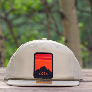 May include: A beige baseball cap featuring a rectangular patch on the front panel. The patch displays a stylized orange and red sunset over a black mountain silhouette with the text C014 printed in orange. A thin olive green cord accents the brim.