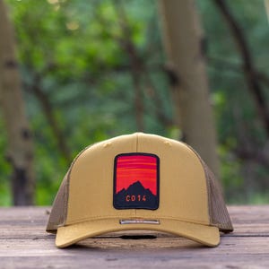 May include: A tan baseball cap with a mesh back and a rectangular patch on the front. The patch features a red and orange sunset graphic above a black mountain silhouette with the text C014 printed in orange at the bottom of the mountain.