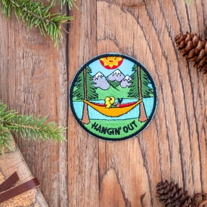 Peut inclure: Écusson rond avec une scène de nature. Une personne se détend dans un hamac jaune entre deux arbres, avec des montagnes et un lac en arrière-plan. Le soleil brille vivement dans le ciel. Le texte "HANGIN' OUT" est en bas.