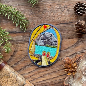 May include: Embroidered patch with a yellow background and a blue border. The patch depicts a mountain range with a lake in the foreground. Two pairs of feet are visible at the bottom of the patch, with a red heart above them.
