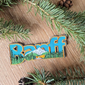 Peut inclure: Un écusson brodé représentant le mot Banff en lettres bleues grasses avec une bordure orange vif. Le motif inclut une scène détaillée d'un sommet montagneux enneigé, une forêt verte dense et un petit canoë jaune sur un lac calme.