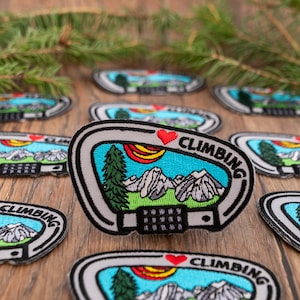 May include: A collection of embroidered patches featuring a mountain scene with a heart and the text "I love climbing". The patches are in various colours, including blue, green, red, and white.