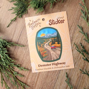 May include: A rectangular vinyl sticker on a light tan backing card with a hole at the top. The sticker features a landscape illustration of the Dempster Highway with a red truck, mountains, and trees. Text reads Balance Explore, Vinyl Sticker, and Dempster Highway, Outdoor Durable & Dishwasher Safe.