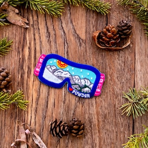 May include: Blue and pink embroidered ski goggle patch with a mountain scene and the word "POWDER" on a wooden background.