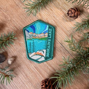 May include: A pentagonal embroidered patch featuring a landscape with a blue lake, rocky mountains, and a green aurora borealis sky. The right side of the patch has a dark green border with the white text MORAINE LAKE embroidered vertically.
