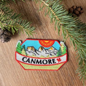 May include: A rectangular embroidered patch with a light blue sky, a bright orange sun, and grey mountain peaks. Green pine trees flank the mountains. A white banner across the bottom features the black text CANMORE and a small red maple leaf.