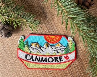 May include: A rectangular embroidered patch with a light blue sky, a bright orange sun, and grey mountain peaks. Green pine trees flank the mountains. A white banner across the bottom features the black text CANMORE and a small red maple leaf.