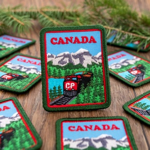 May include: Embroidered patch depicting a Canadian landscape with a red train marked "CP" on tracks, mountains, and the word "CANADA" in red. The patch has a green border, set against a wooden surface with pine needles.