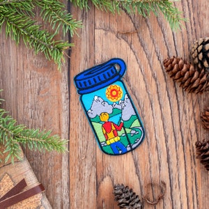 May include: An embroidered patch shaped like a jar, featuring a hiker in a red shirt and a mountain scene. The patch is blue and green with a white border and is displayed on a wooden surface.