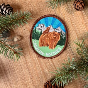 May include: A circular embroidered patch featuring a brown bear standing in front of green pine trees and white snow-capped mountains against a blue sky with sun rays. The text CANADIAN ROCKIES is stitched in white along the bottom edge.