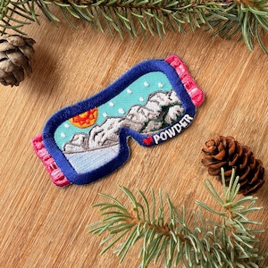 May include: An embroidered patch shaped like ski goggles with a blue frame and pink strap ends. The lens features a mountain landscape with a yellow sun, white snow dots, and the word POWDER in white text next to a small red heart icon.