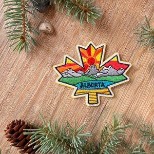 May include: A maple leaf shaped embroidered patch featuring a sunrise over grey mountains, green hills, and a blue lake. The word ALBERTA is stitched in black across the centre. The design includes yellow, orange, red, green, blue, and grey thread.