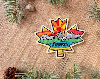May include: A maple leaf shaped embroidered patch featuring a sunrise over grey mountains, green hills, and a blue lake. The word ALBERTA is stitched in black across the centre. The design includes yellow, orange, red, green, blue, and grey thread.