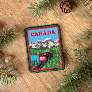 May include: A rectangular embroidered patch featuring a red train engine labeled CP traveling through a green forest with snow-capped mountains in the background and the word CANADA printed in red at the top, all bordered by a thick red frame.