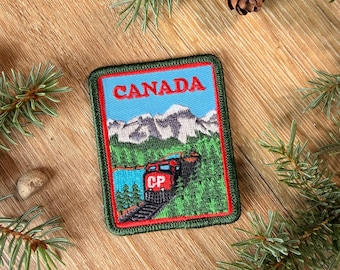 May include: A rectangular embroidered patch featuring a red train engine labelled CP travelling through a green forest with snow-capped mountains in the background and the word CANADA printed in red at the top, all bordered by a thick red frame.