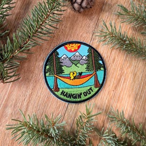 Op de afbeelding: Een ronde geborduurde patch met een landschapsscène met een heldere gele zon, grijze bergen, groene dennenbomen en een blauw meer. Een oranje hangmat hangt tussen twee bomen met de tekst HANGIN OUT in zwarte stiksels aan de onderkant.