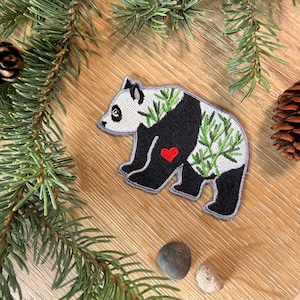 May include: An embroidered patch shaped like a panda bear with a white face, black ears, and black limbs. The body features a green bamboo leaf pattern on a white background, a solid black midsection, and a small, bright red embroidered heart.