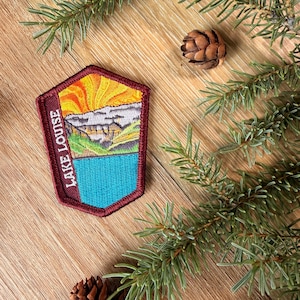 Op de afbeelding: Een vijfhoekige geborduurde patch met een landschap van Lake Louise, inclusief een heldergele zon, grijze bergen, groene hellingen en een turquoise meer. De tekst "LAKE LOUISE" is verticaal in het wit geborduurd op een donkerrode rand.