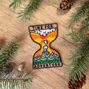 May include: An embroidered patch shaped like an hourglass with the text TIME FOR ADVENTURE. The design features a sun, clouds, grey mountains, and green pine trees in orange, yellow, white, and green thread on a black background border.