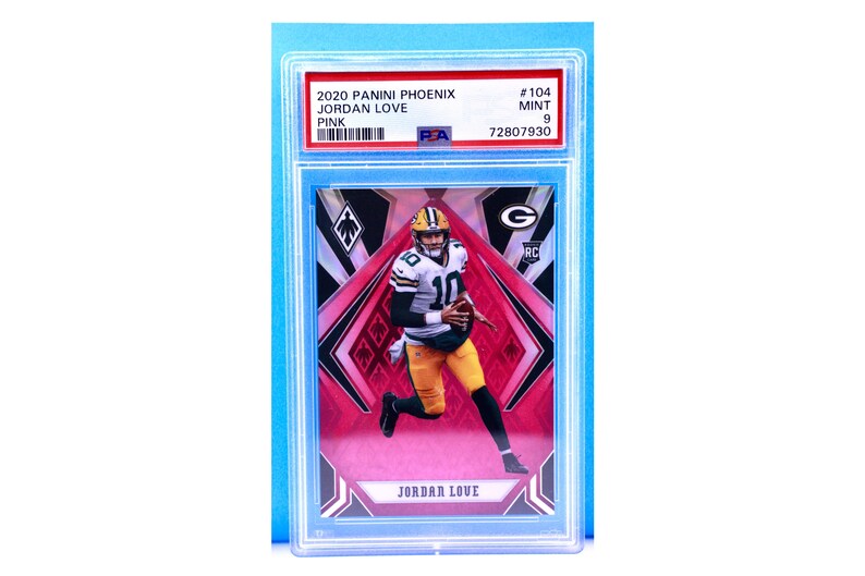 May include: A graded trading card of Jordan Love, a football player for the Green Bay Packers. The card is from the 2020 Panini Phoenix set and is a pink parallel. The card is graded a 9 by PSA and has a black and red design with a diamond pattern.