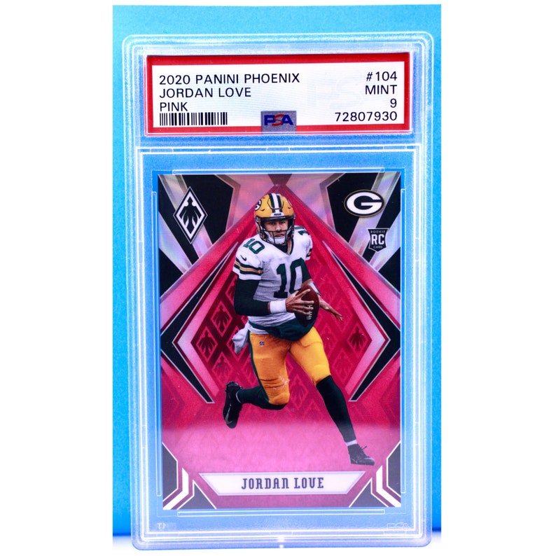 May include: A graded trading card of Jordan Love, a football player for the Green Bay Packers. The card is from the 2020 Panini Phoenix set and is a rookie card. The card is graded a 9 by PSA and has a pink background.