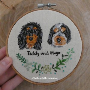 May include: Embroidered portrait of two dogs, Teddy and Hugo, with green foliage surrounding their names. The embroidery is framed in a wooden hoop and the artist's Instagram handle, @bethanyduffyembroidery, is included below.