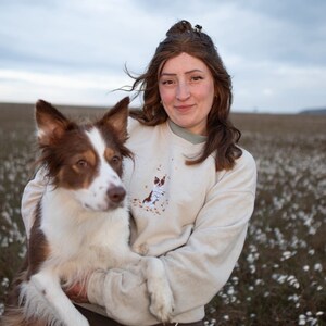 May include: A person wearing a cream sweatshirt with a dog graphic holds a brown and white dog. The dog has brown ears and a white muzzle. The background is a field with white flowers under a cloudy sky.