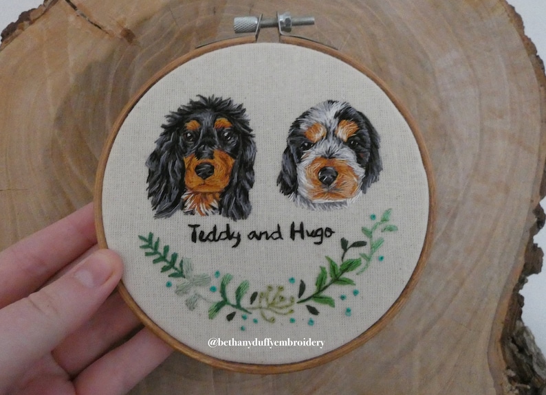 May include: Embroidered portrait of two dogs, Teddy and Hugo, with green foliage surrounding their names. The embroidery is framed in a wooden hoop and the artist's Instagram handle, @bethanyduffyembroidery, is included below.