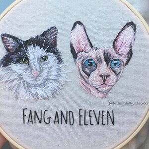 May include: Embroidered portraits of two cats, one black and white and one gray, with the names "Fang and Eleven" stitched below. The embroidery is on a round, beige canvas.