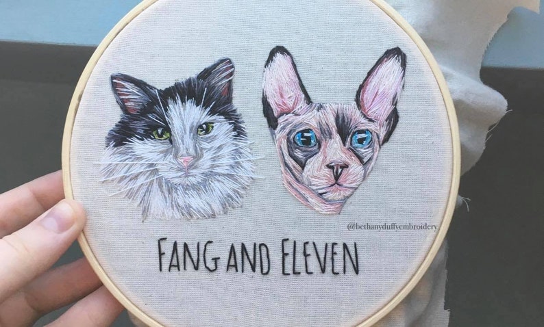 May include: Embroidered portraits of two cats, one black and white and one gray, with the names "Fang and Eleven" stitched below. The embroidery is on a round, beige canvas.