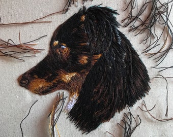 Puede incluir: Retrato bordado de la cabeza de un perro de perfil. El perro tiene pelaje negro y marrón, con puntadas detalladas que crean textura y profundidad. El fondo es una tela beige claro, con hilos sueltos de varios colores.