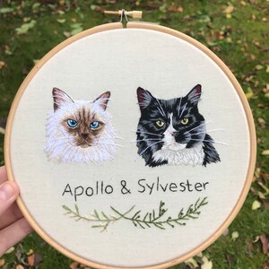 May include: Embroidered hoop art featuring two cats, one white with blue eyes and one black and white, with the text "Apollo & Sylvester" and a green sprig of rosemary below.
