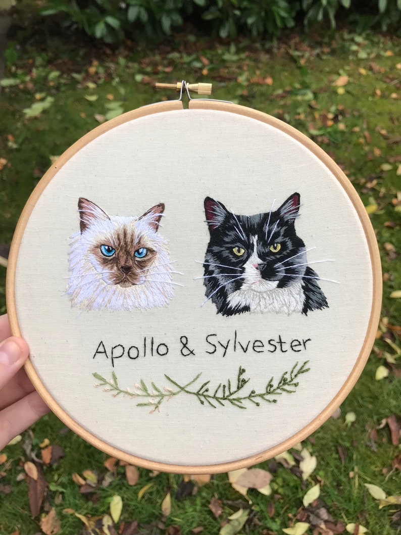 May include: Embroidered hoop art featuring two cats, one white with blue eyes and one black and white, with the text "Apollo & Sylvester" and a green sprig of rosemary below.