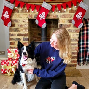 May include: A festive scene with a Christmas theme. A woman kneels beside a black and white dog, both appearing happy. A navy blue sweatshirt with a Santa design is visible. Red and white wrapped gifts and Christmas stockings decorate the fireplace.