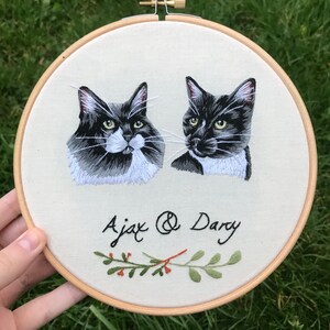 May include: Embroidered hoop art featuring two black and white cats with green foliage and red berries. The cats are named Ajax and Dary.