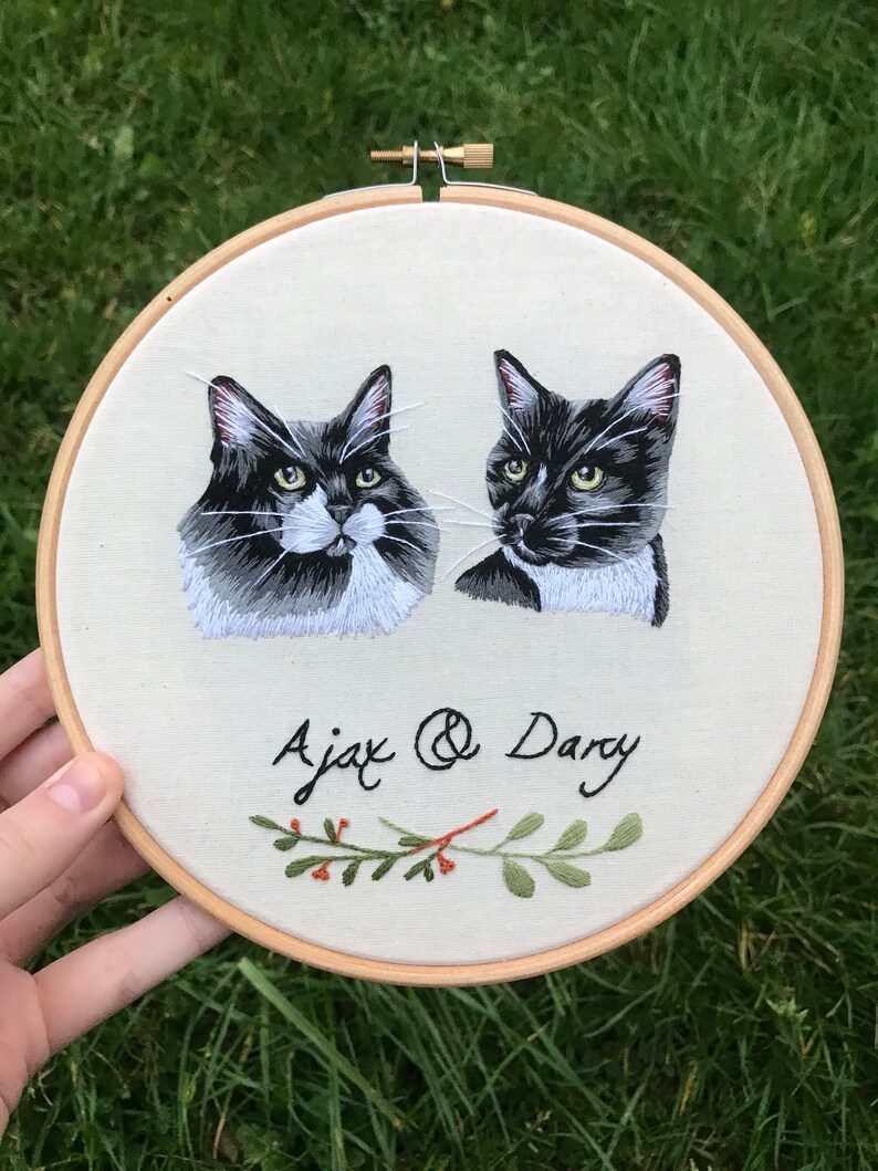 May include: Embroidered hoop art featuring two black and white cats with green foliage and red berries. The cats are named Ajax and Dary.