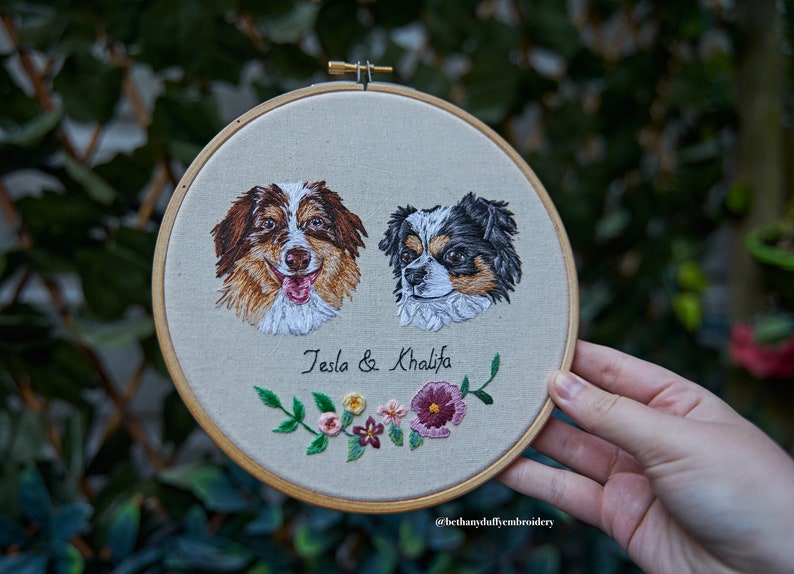 May include: Embroidered portrait of two dogs, Tesla and Khalifa, with a floral design below their names. The embroidery is framed in a wooden hoop.