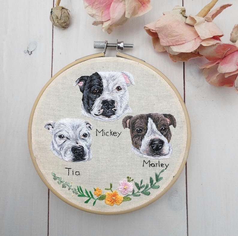 May include: Embroidered hoop art featuring three dogs, a black and white dog named Mickey, a white dog named Tia, and a brown and white dog named Marley. The hoop is decorated with a floral border.