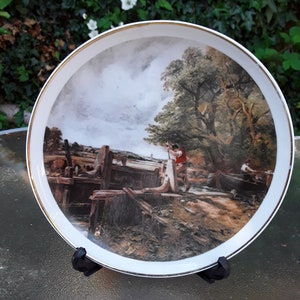 May include: A white porcelain plate with a gold rim features a painting of a canal scene with two figures in the foreground. The scene depicts a wooden lock with a boat passing through. The sky is cloudy and the trees are green.