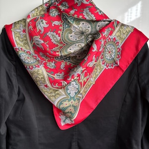 May include: A square scarf featuring a vibrant red background with an intricate paisley pattern in shades of olive green, white, and black. The scarf is draped over a solid black blazer, showcasing a detailed border design with floral motifs.