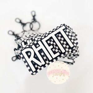 May include: A black and white checkered keychain with the name "Rhett" in white letters. The keychain is attached to a silver key ring. The keychain is made by Pebble Hill Customs.
