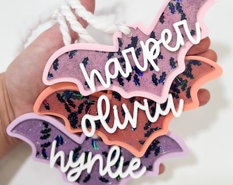 May include: Three layered bat ornaments with glitter and a white rope. The bats are pink, orange, and purple with the names "Harper", "Olivia", and "Hyndlie" written in white.