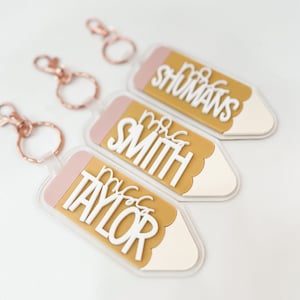 May include: Three gold pencil-shaped keychains with pink accents and white lettering. The keychains read "Mrs. Taylor", "Mrs. Smith", and "Mrs. Shomans".